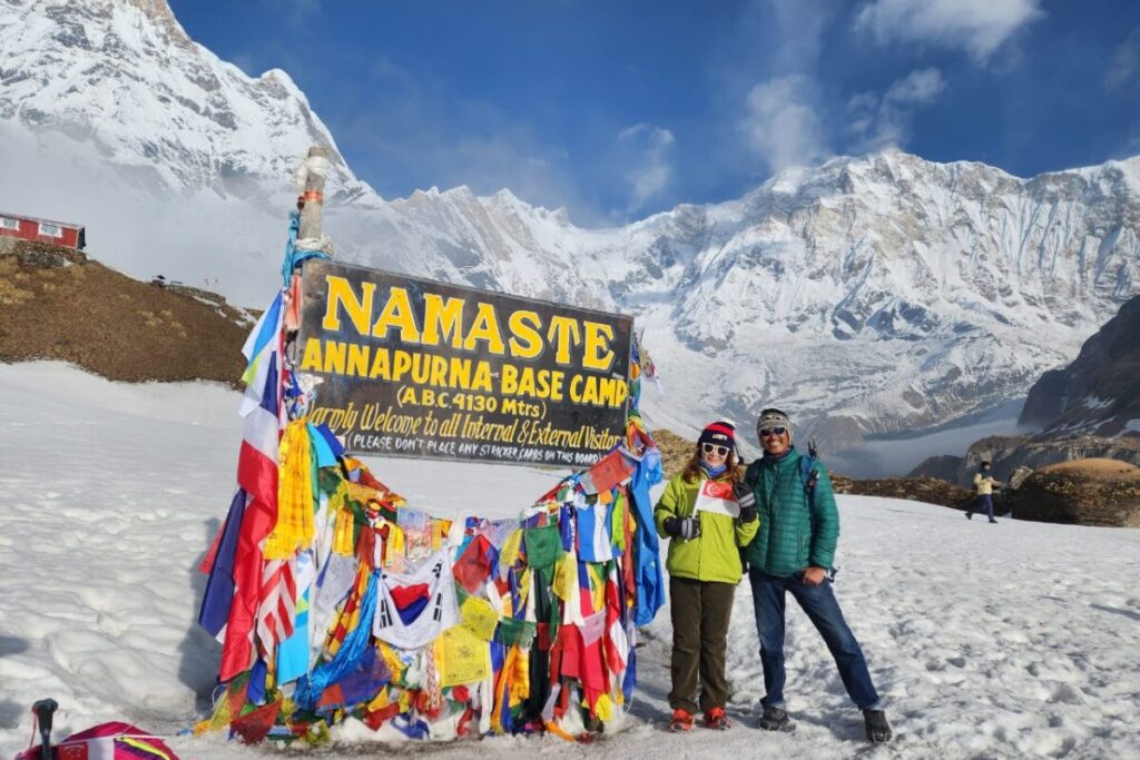Two happy trekkers standing by the Namaste Annapurna Base Camp sign at 4,130m altitude surrounded by prayer flags.