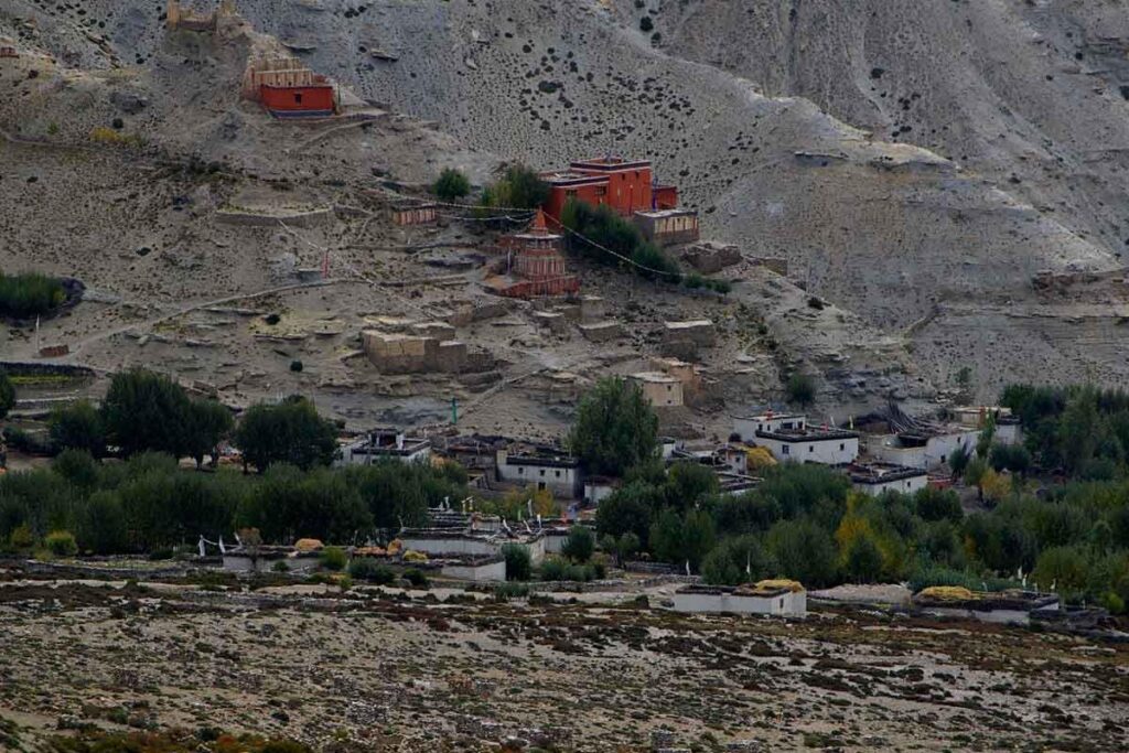 Scenic view of Ghiling village in Upper Mustang along the Trek to Lo Manthang, featuring a red Buddhist monastery, temple, brown hills, and prayer flags.