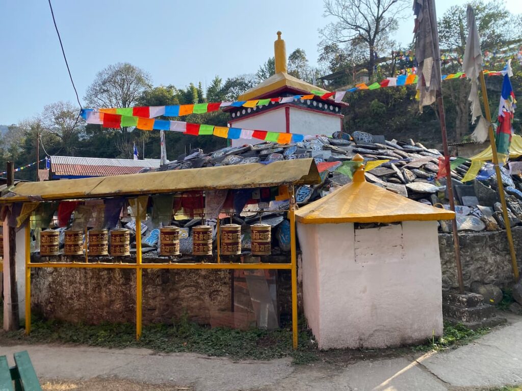 Row of golden Tibetan prayer wheels beside a pile of carved mani stones and colourful prayer flags at a Tibetan refugee settlement in Pokhara