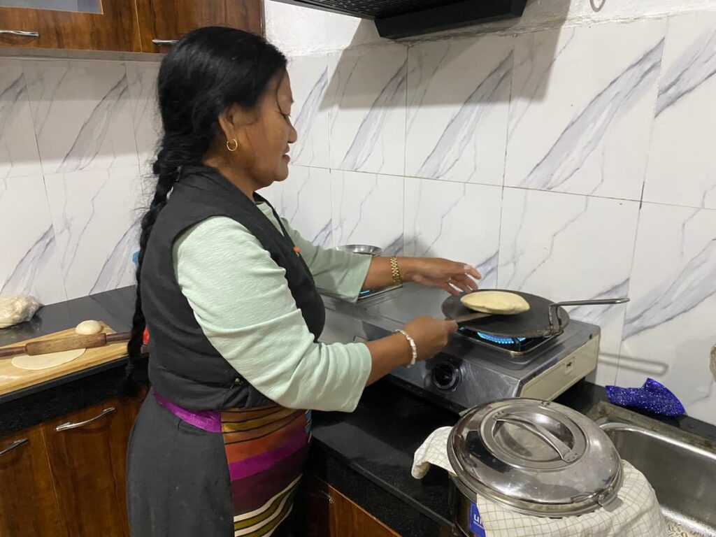 A Tibetan woman in traditional dress cooking homemade flatbread on a stove in a local family home — a highlight of the afternoon cultural tour in Pokhara