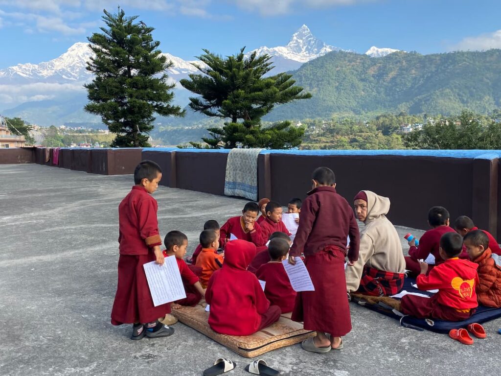 Young novice monks in red robes reciting scriptures on a monastery rooftop with snow-capped Annapurna mountains in the background