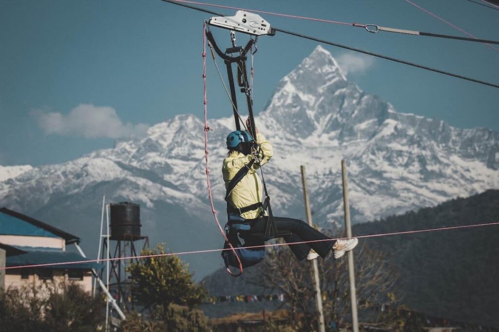 Zipline rider racing above green hills with Machhapuchhre Fishtail peak in the background.