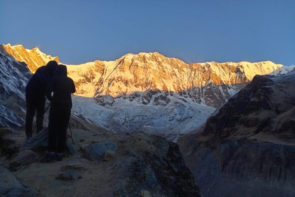 Two hikers silhouetted at sunrise watching golden light on a Himalayan glacier
