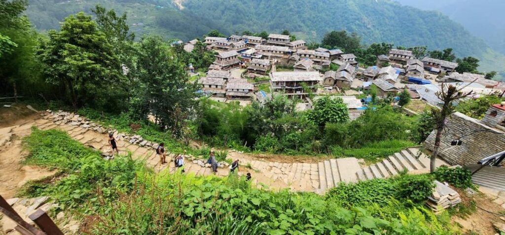 Bird's-eye view of a traditional stone-roofed Himalayan village nestled among green terraced hillsides with hikers walking up a stone path.