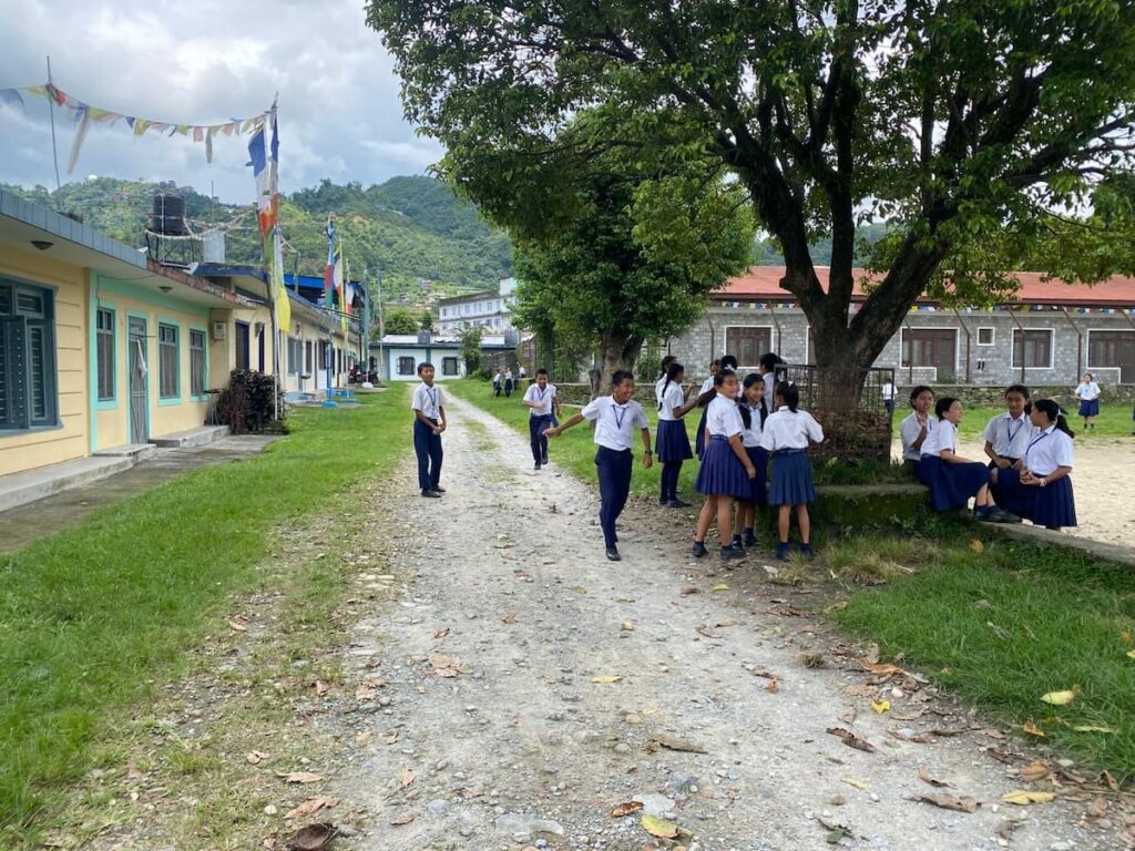 School children near a tree inside the Tibetan Refugee Settlement in Pokhara with Buddhist prayer flags along the pathway — a cultural stop on the Pokhara sightseeing trip